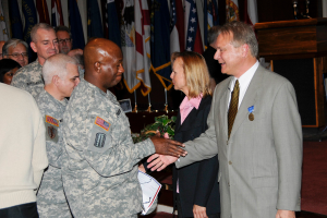 Eine Gruppe von Menschen, die mit zwei Personen Hand gibt, Flaggen im Hintergrund, ein Podium mit Mikrofon und eine Topfpflanze in der Nähe.
