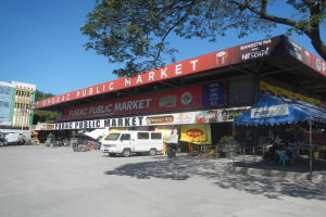Ein öffentlicher Markt mit einem vor ihm geparkten Van, umgeben von Gebäuden, Geschäften, Namensschildern, Kraftfahrzeugen auf der Straße, Menschen unter einem Zelt sitzend, Geländern, Bäumen und einem klaren blauen Himmel.