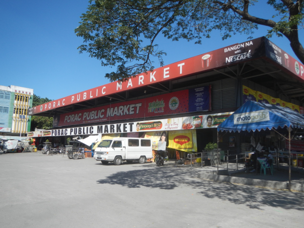Ein öffentlicher Markt mit einem vor ihm geparkten Van, umgeben von Gebäuden, Geschäften, Namensschildern, Kraftfahrzeugen auf der Straße, Menschen unter einem Zelt sitzend, Geländern, Bäumen und einem klaren blauen Himmel.