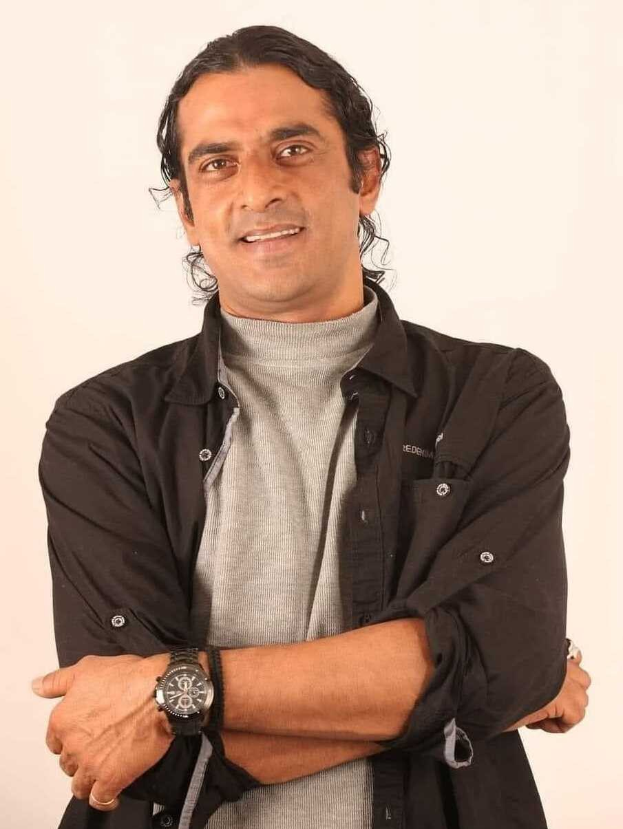 Ein Mann in einem schwarzen und aschfarbenen Kleid mit verschränkten Armen, der eine Uhr trägt und vor einem weißen Hintergrund steht, in Gedanken versunken.