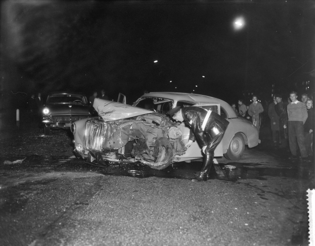 Ein Schwarz-Weiß-Foto eines auf der Straße liegenden Wagens, umgeben von mehreren Personen und beleuchtet von Hintergrundlichtern.