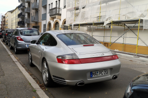 Ein silberner Porsche steht auf einer Straße, umgeben von Gebäuden mit Fenstern, Geländern und Balkonen, mit einem im Bau befindlichen Gebäude auf der rechten Seite und dem Himmel im Hintergrund.