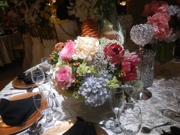 Ein formeller Esstisch mit Tellern, Besteck, Gläsern, Servietten und Blumenvasen, der eine florale Tischdeko und im Hintergrund Stühle und andere Tische zeigt.