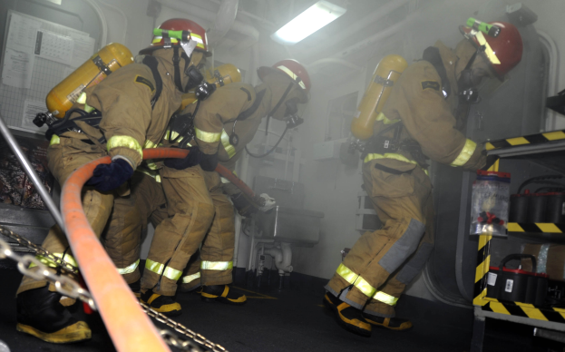 Feuerwehrleute in Schutzausrüstung arbeiten in der Nähe eines Feuerwehrautos mit einem Gestell von Gegenständen und Hintergrundrohren und -lichtern.