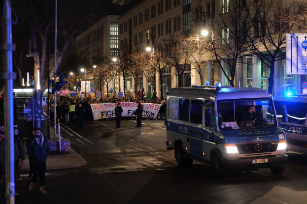 Eine Gruppe von Menschen, die nachts eine Straße entlanggehen, mit einem Polizeiwagen auf der Seite und einer sichtbaren Fahne im Hintergrund.