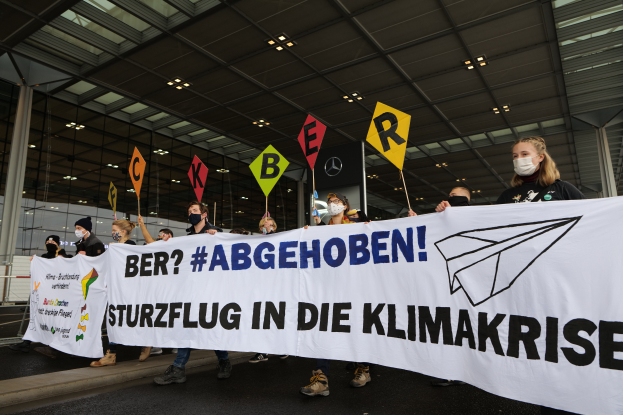 Eine Gruppe maskierter Individuals mit einem Banner "Sturzflug in die Klimakrise" vor einem Glasgebäude mit Säulen, beleuchtet von Deckenlampen.