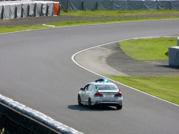 Polizeiauto auf einer Rennstrecke mit Gras auf beiden Seiten, Menschen, Zelte und andere Gegenstände im Hintergrund sichtbar.