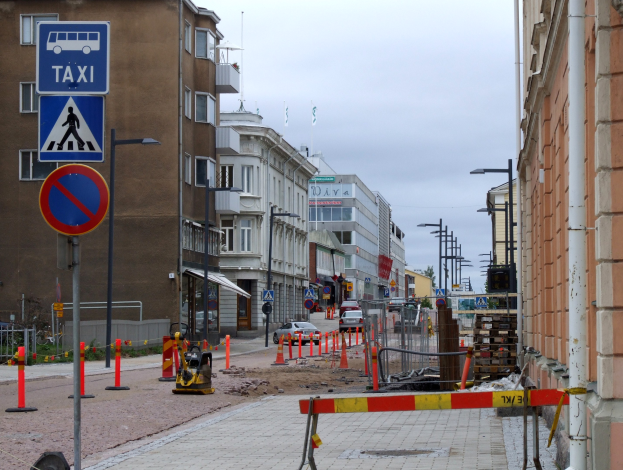 Stadtstraße mit Gebäuden, Straßnpfählen, Lichtern, Schildern, Verkehrskegeln, Fahrzeugen, Absperrpölen, Bäumen, einer Baustelle mit vielen Verkehrsschildern und einem bewölktem Himmel.