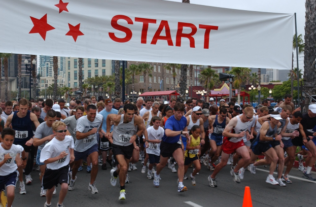 Gruppe von Läufern bei einem Marathon an einer Verkehrsinsel vorbei, mit einem Banner und Gebäuden im Hintergrund bei klarem blauem Himmel.