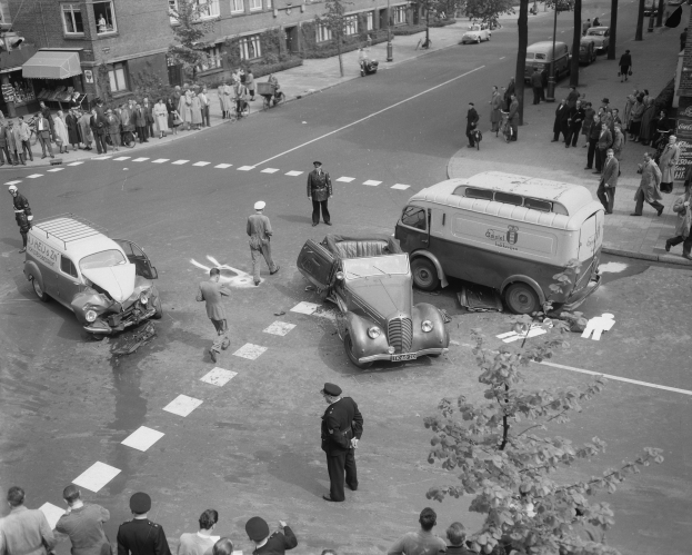 Schwarz-weiß-Szene eines Autounfalls an einer Straßenkreuzung mit einem Lieferwagen, anderen Fahrzeugen und Menschen in der Nähe; Gebäude, Bäume, Pfosten und Schilder im Hintergrund.