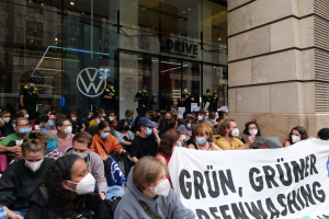 Eine große Gruppe von Menschen mit Masken und einem Banner versammelt sich vor einem Glasgebäude mit Logo und Text und demonstriert in Berlin, Deutschland für den Abzug der Volkswagen AG.