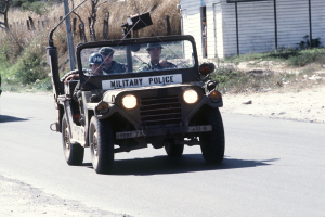 Ein Militärpolizeijeep mit Helmträgern fährt eine Straße neben einem Gebäude entlang, mit einem Haus auf der rechten Seite und Bäumen im Hintergrund.