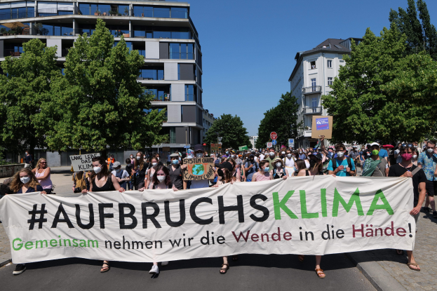 Eine Gruppe von Menschen mit Masken, die ein Banner mit der Aufschrift 'Aufbruchsklima' vor einem Gebäude halten, umgeben von Bäumen und einem klaren Himmel.