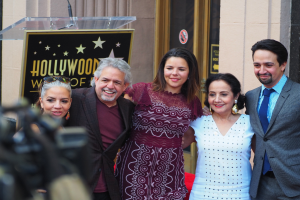 Eine Gruppe von Menschen steht vor einem Hollywood Walk of Fame-Star, in der Mitte ein Podium und ein Mikrofon, ein Schild mit Text dahinter und links eine Person mit einer Kamera, Wände und eine Tür im Hintergrund.