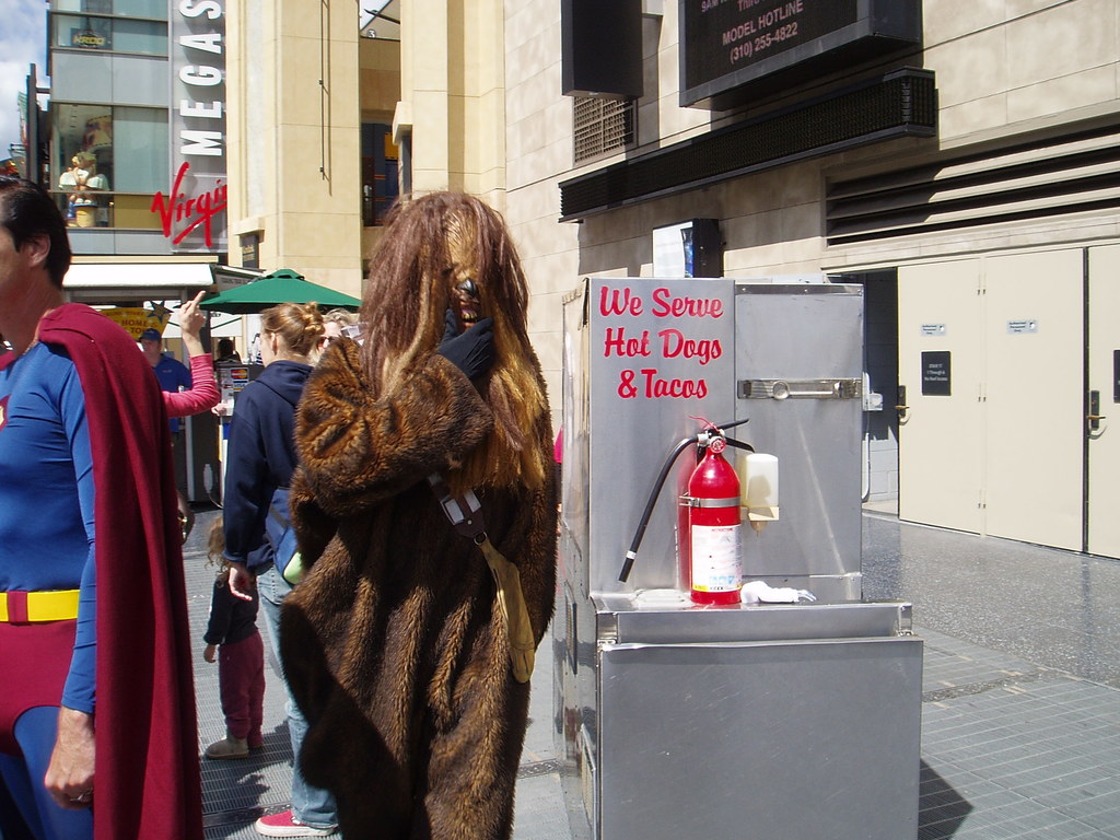 Eine Gruppe von Menschen, einige in Kostümen, versammelt sich vor Gebäuden mit sichtbarer Beschriftung, einer Box mit Schrift und einem Feuerlöscher.