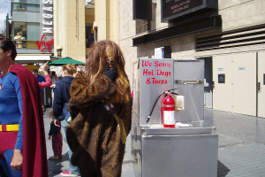 Eine Gruppe von Menschen, einige in Kostümen, versammelt sich vor Gebäuden mit sichtbarer Beschriftung, einer Box mit Schrift und einem Feuerlöscher.