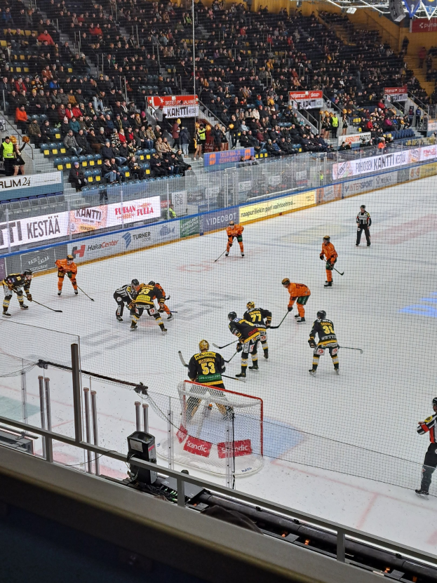 Gruppe von Menschen, die Hockey auf einer Indoor-Eisfläche mit einem Netz und einer Umrandung spielen, mit Zuschauern im Hintergrund, sichtbaren Bannern und Überdachungsbeleuchtung.