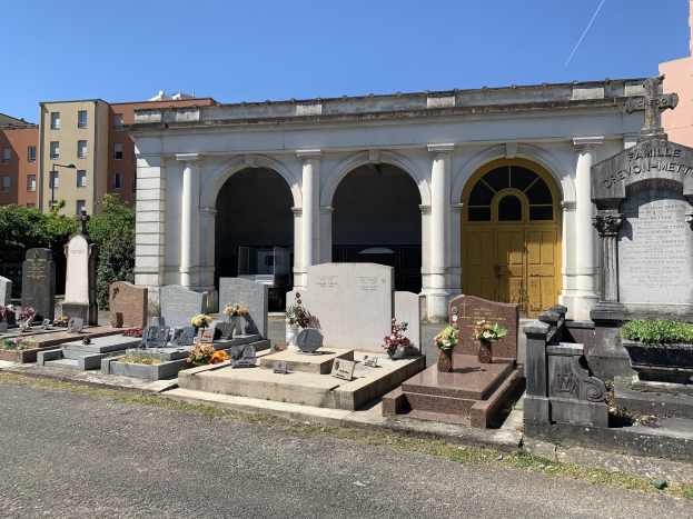 Friedhof mit zahlreichen Grabsteinen und Blumen, gelbgetürmtem Gebäude, Bäumen, benachbarten Gebäuden mit Fenstern und einem klaren blauen Himmel.