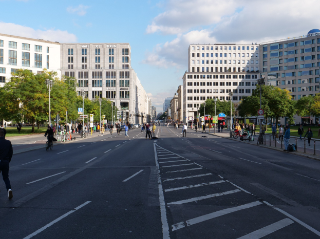 Eine belebte Stadtstraße in Berlin, Deutschland, mit Fußgängern und Radfahrern, hohen Gebäuden, Bäumen, Laternenmasten und Schildern unter einem bewölkten Himmel.