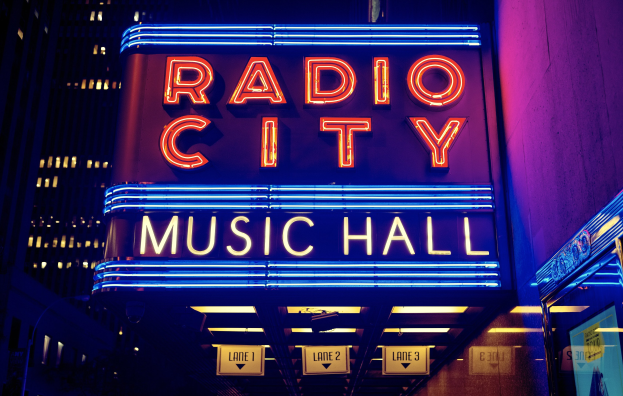 Außenansicht des Radio City Music Hall in New York City bei Nacht mit heller Beleuchtung und hohen umliegenden Gebäuden, mit einem "Radio City"-Schild und zusätzlichen Schildern auf der rechten Seite.