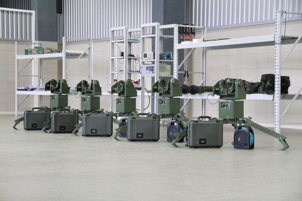 Gruppe von Militärrobotern auf einem Boden mit Koffern und Objekten auf Gestellen, vor einer Wand mit Geländer im Hintergrund.