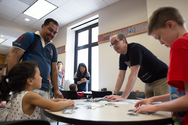 Eine Gruppe von Menschen sitzt um einen Tisch und spielt ein Spiel mit Puzzles darauf, in einem Raum mit einem Fenster, einer Tafel und Deckenlampen.