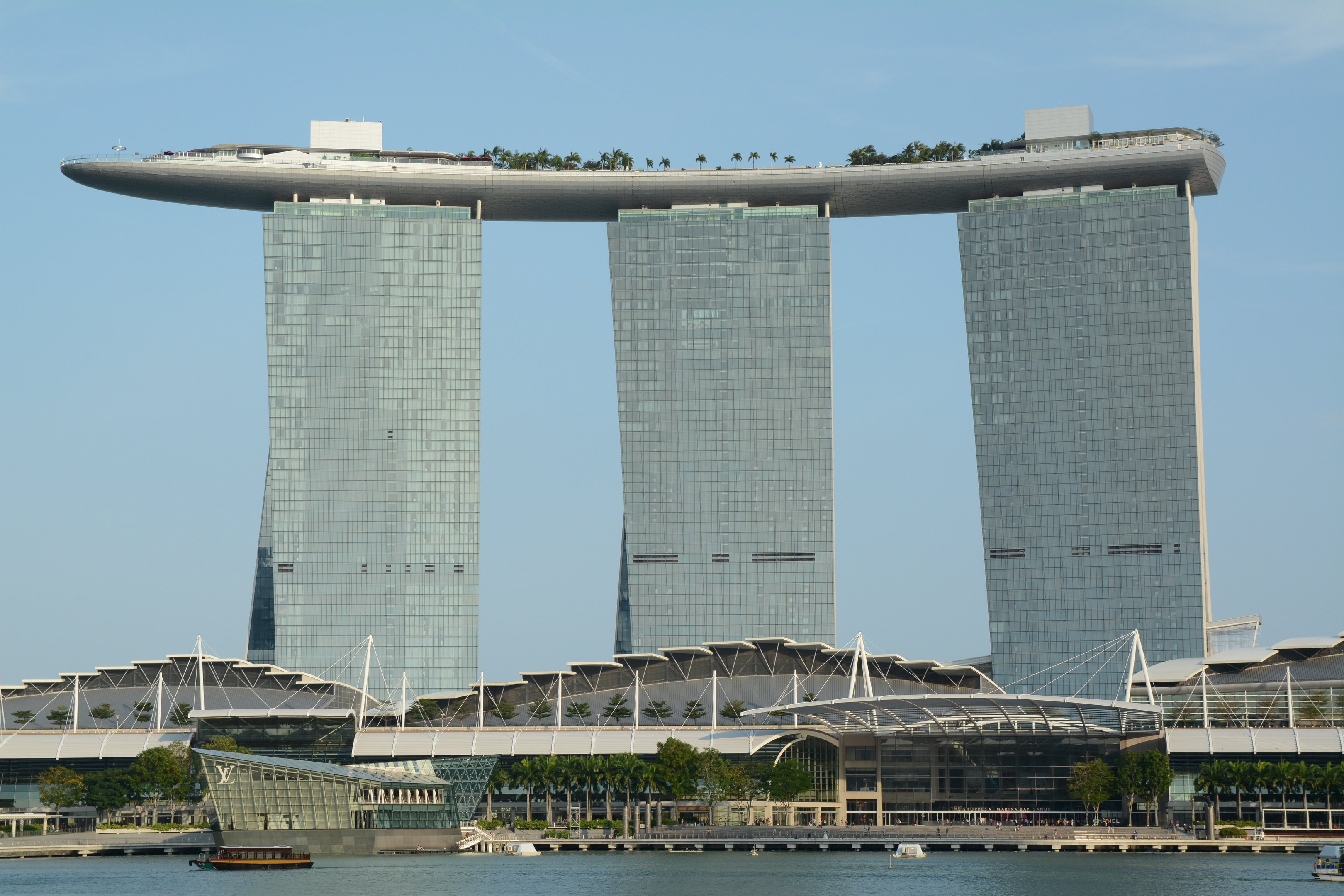 Das ikonische Marina Bay Sands Hotel in Singapur mit seinen drei turmhohen Wolkenkratzern, die von einem Dachgarten verbunden sind, umgeben von üppiger Vegetation und Booten auf dem Wasser darunter, unter einem bewölkten Himmel.