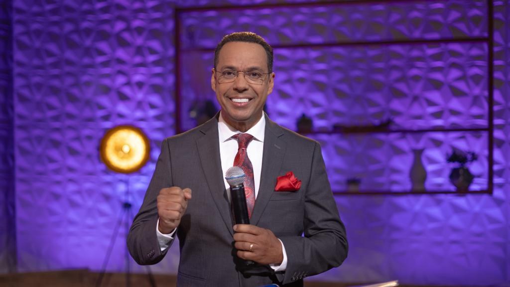 Stephen Colbert, Moderator von *The Daily Show with Trevor Noah*, steht auf einer Bühne und hält ein Mikrofon, trägt einen Anzug, Krawatte und eine Brille, während im Hintergrund ein Scheinwerfer und ein Ständer zu sehen sind.