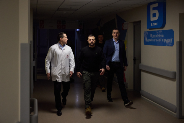 Eine Gruppe von Männern geht einen Krankenhausflur entlang mit Deckenleuchten, einer Tür an der rechten Wand und Texttafeln an der linken Wand, die besorgt aussehen.