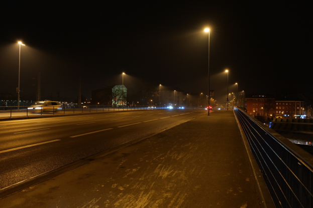 Langzeitaufnahme einer nächtlichen Stadtstraße mit fahrenden Fahrzeugen, die Lichtspuren erzeugen, sichtbare Geländer, Laternen, Gebäude, Bäume und einen dunklen Himmel.
