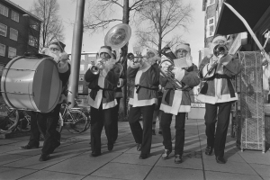 Eine Gruppe von Männern in Santa-Klaus-Kostümen, die auf einem Gehweg Musikinstrumente spielen, umgeben von Fahrrädern, Pfählen, Bäumen, Gebäuden mit Fenstern, unter einem klaren Himmel, dargestellt in Schwarz-Weiß.
