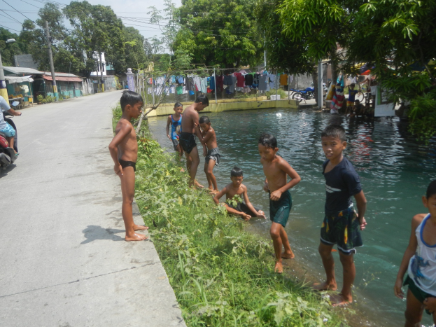 Eine Gruppe von Kindern spielt in flachem Wasser neben einer Straße, einige stehen und andere sitzen auf Gras, mit einer Person auf einem Motorrad links und Häusern, Bäumen und einem klaren blauen Himmel im Hintergrund.