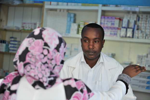 Eine Apothekerin in einem weißen Laborkittel steht vor einer sitzenden Frau in einer Apotheke, mit Regalen voller Boxen und Gegenständen im Hintergrund.
