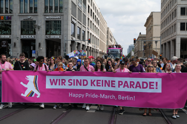 Eine Gruppe von Menschen, die eine Straße in Berlin, Deutschland, entlanggehen und ein pinkfarbenes Banner mit der Aufschrift "Happy Pride March" halten, mit Gebäuden, Laternenmasten und Verkehrszeichen, die die Straße säumen, unter einem bewölkten Himmel.