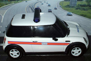 Ein weißes Polizeiauto steht im Vordergrund mit einer belebten Straße voller fahrender Autos dahinter, flankiert von Gras und Bäumen auf beiden Seiten, in einer ländlichen Umgebung.