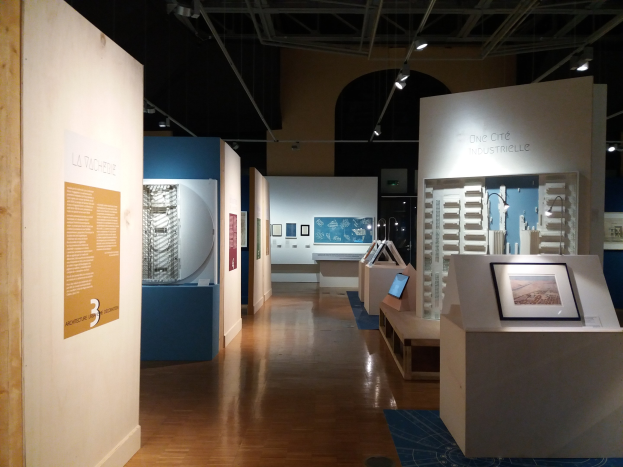 Ein Raum mit verschiedenen Kunstwerken, darunter gerahmte Fotos auf Tischen, Texttafeln an den Wänden, Deckenleuchten und einem Teppichboden, was auf eine Ausstellung im Museum of Modern Art hindeutet.
