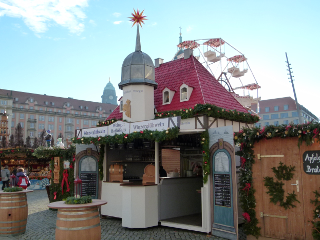 Kleines Gebäude mit rotem Dach und einem Riesenrad davor, umgeben von festlichen Oktoberfest-Dekorationen, Menschen und Gegenständen, vor einem Hintergrund aus Gebäuden, Bäumen und einem bewölkten Himmel.