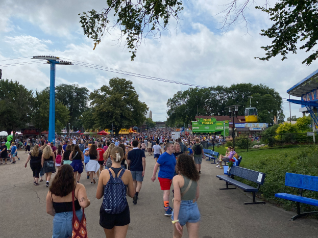 Eine Menschenmenge, die eine Straße mit Bänken, Pflanzen, Gras, Bäumen, Pfählen, Lampen, Kabeln und einer Hütte entlanggeht, wobei einige Taschen tragen, unter einem bewölkten Himmel.