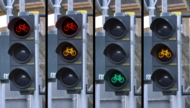 Verkehrsampel mit Fahrradsymbol, an einem Mast mit zusätzlichen Masten und einem Gebäude im Hintergrund angebracht.