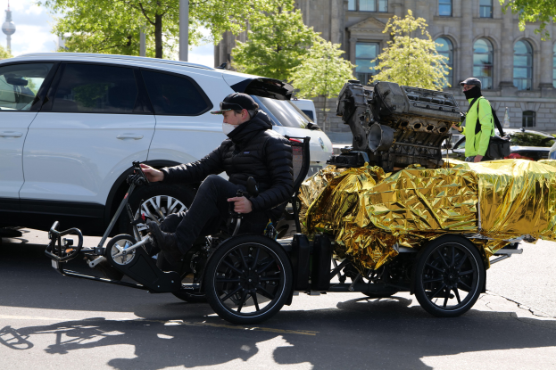 Ein Mann im Rollstuhl mit einem großen Motor auf dem Rücken, umgeben von Fahrzeugen auf einer Straße mit Bäumen, Gebäuden, Masten und einem klaren blauen Himmel im Hintergrund; er trägt eine schwarze Jacke, eine Kappe und hält ein Objekt in der Hand.