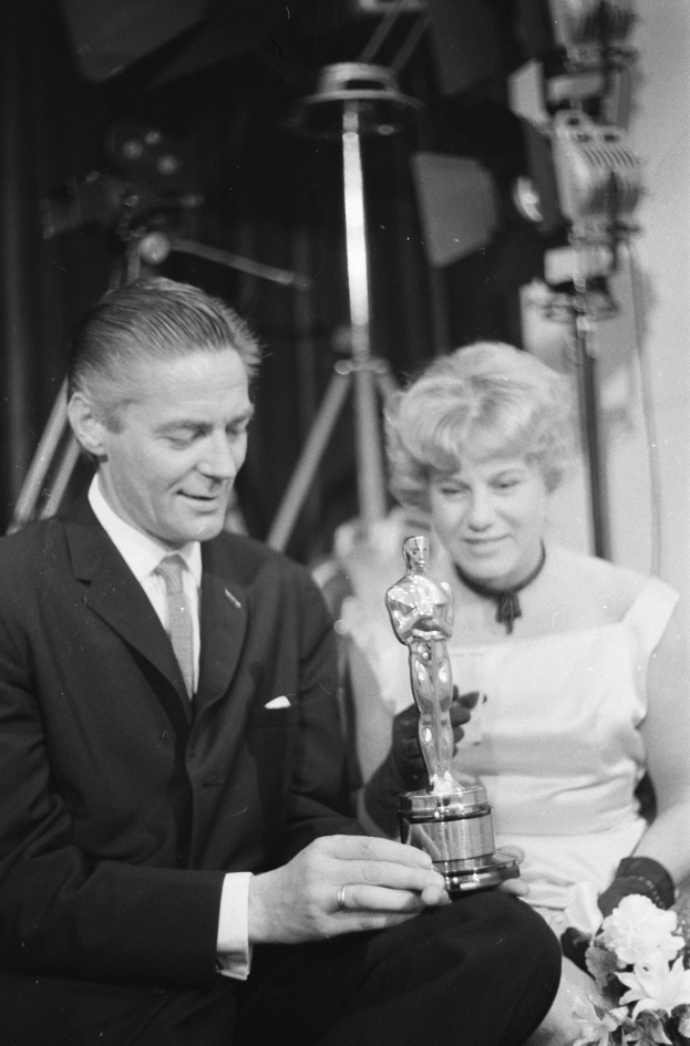 Ein Mann in einem schwarzen Blazer, weißem Hemd und Krawatte hölt eine Oscar-Statue, während er neben einer Frau in Abendgarderobe sitzt, die einen Blumenstrauß hölt, mit Lichtern im Hintergrund.