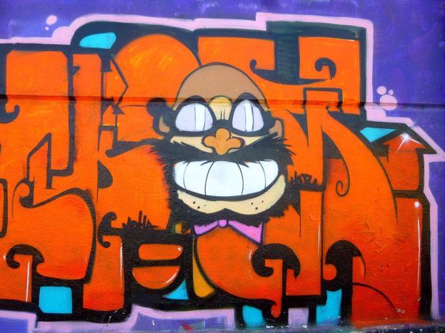 Graffiti-Gemälde eines Cartoon-Charakters mit großen Augen, einem breiten Grinsen und einem verschmitzten Ausdruck auf einem Gebäude, mit leuchtenden Farben und auffälliger Begleittext.