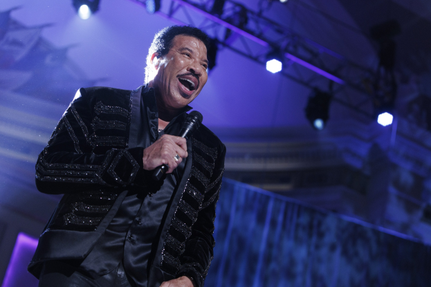 Lionel Richie performs on stage at the Grammys, wearing a black dress and holding a microphone, with bright stage lights creating a dramatic atmosphere.