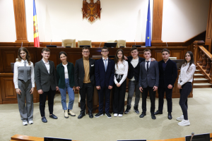Gruppe von strahlenden Menschen, die vor einem Gerichtssaal mit Monitoren und Mikrofonen auf Tischen, Holzplattform mit Fahnen und Stühlen im Hintergrund und Treppe auf der rechten Seite posieren.