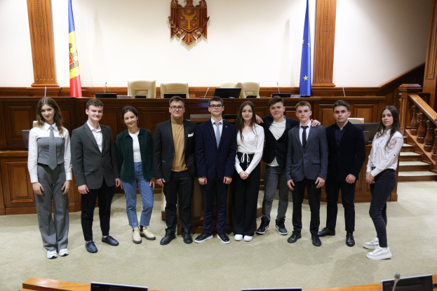 Gruppe von strahlenden Menschen, die vor einem Gerichtssaal mit Monitoren und Mikrofonen auf Tischen, Holzplattform mit Fahnen und Stühlen im Hintergrund und Treppe auf der rechten Seite posieren.