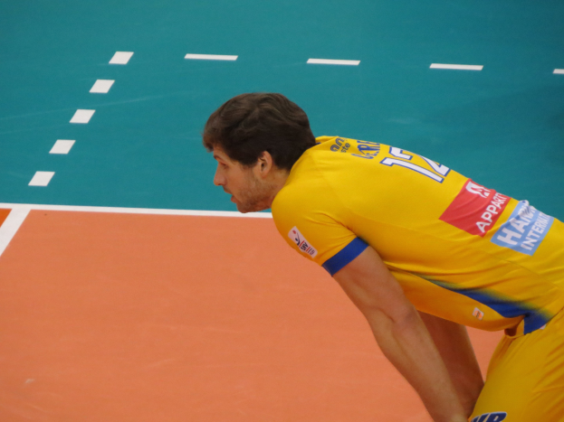 Ein Mann in einem gelben Hemd steht auf einem Volleyballfeld und bereitet sich auf den Aufschlag vor, trägt ein T-Shirt mit der Aufschrift "Volleyball".