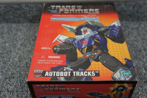 Ein roter, oranger und blauer Kasten mit einem Roboter und dem Text "AUTOBOT TRACKS" ├Âber dem Kasten, vor einem unscharfen schwarzen Hintergrund.