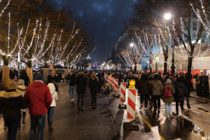 Eine Menschenmenge, die nachts eine Straße entlanggeht, geschmückt mit Weihnachtslichtern an Bäumen und Gebäuden, unter einem bewölkten Himmel.
