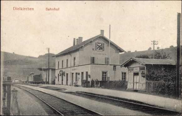 Schwarzes und weißes Foto des Bahnhofs Dielkirchen in Deutschland mit einem Zug auf den Schienen, umgeben von Häusern, Bäumen und Menschen, mit Hügeln und einem klaren Himmel im Hintergrund.