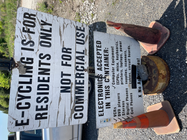 Ein Schild mit der Aufschrift "E-Bike-Fahren nur für Anwohner" steht an der Straßenseite, umgeben von Verkehrskegeln und anderen Gegenständen, mit einem Fahrzeug, Bäumen und einem klaren blauen Himmel im Hintergrund.